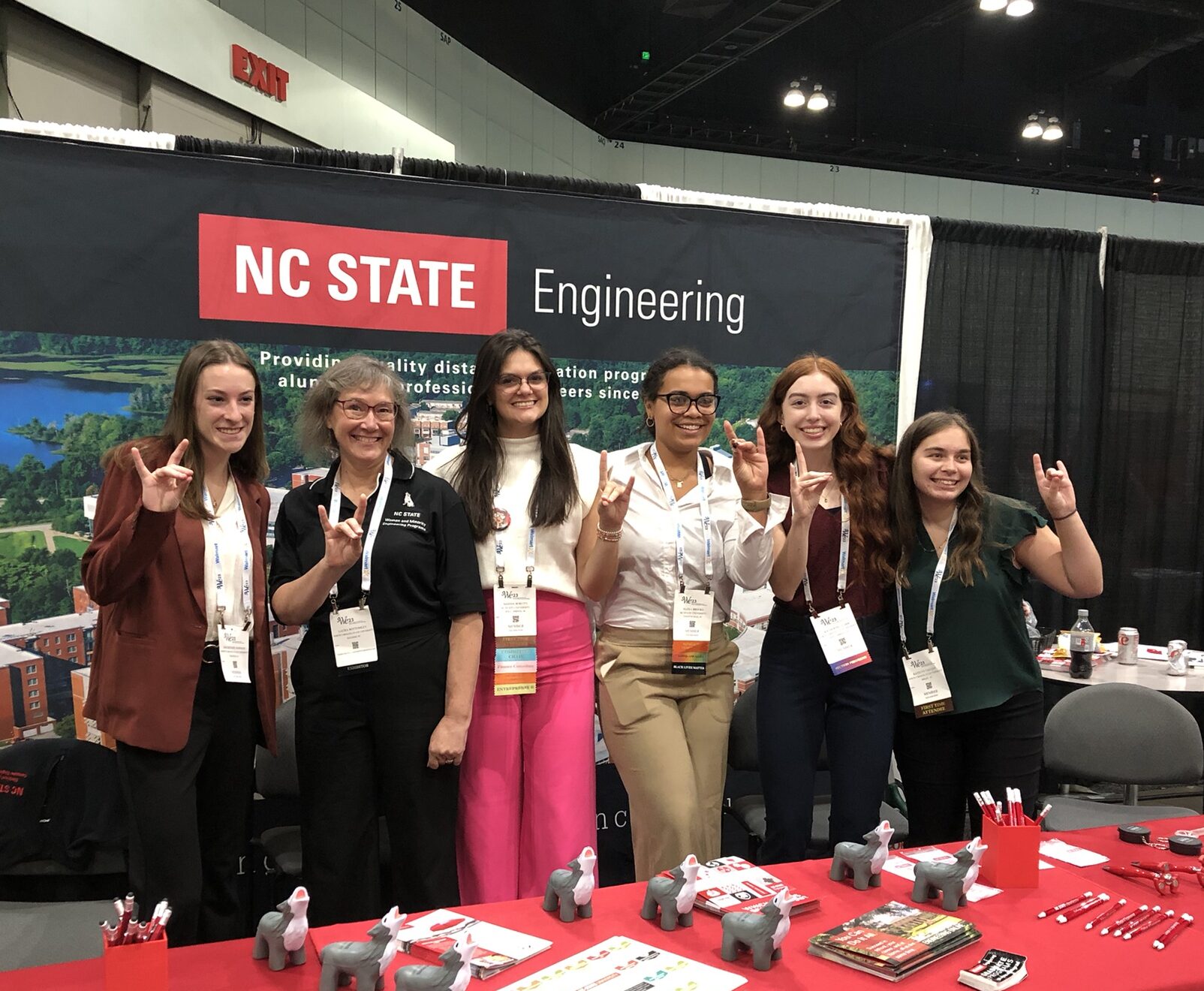Six women wearing conference attire and badges stand in front of an NC State Engineering banner, all holding up the wolfpack hand sign.