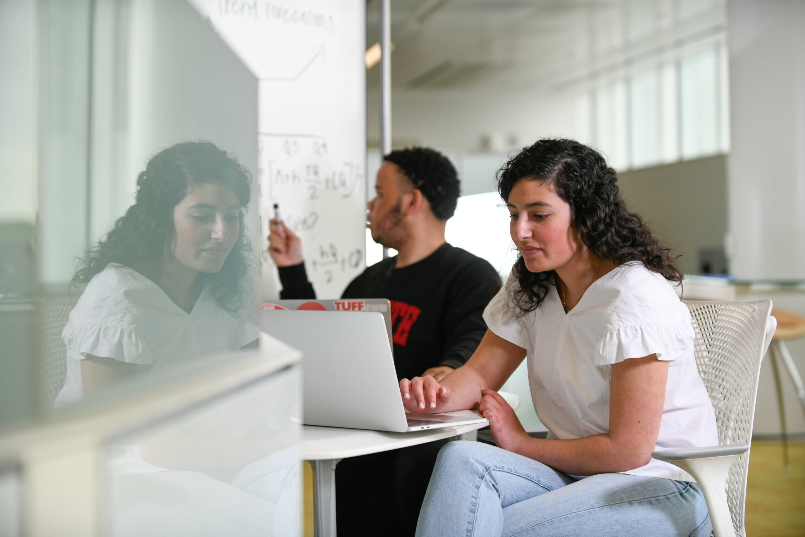 Two students sit in front of a whiteboard with text written on it. Once student points to the board while the other looks at the laptop sitting in front of them. 