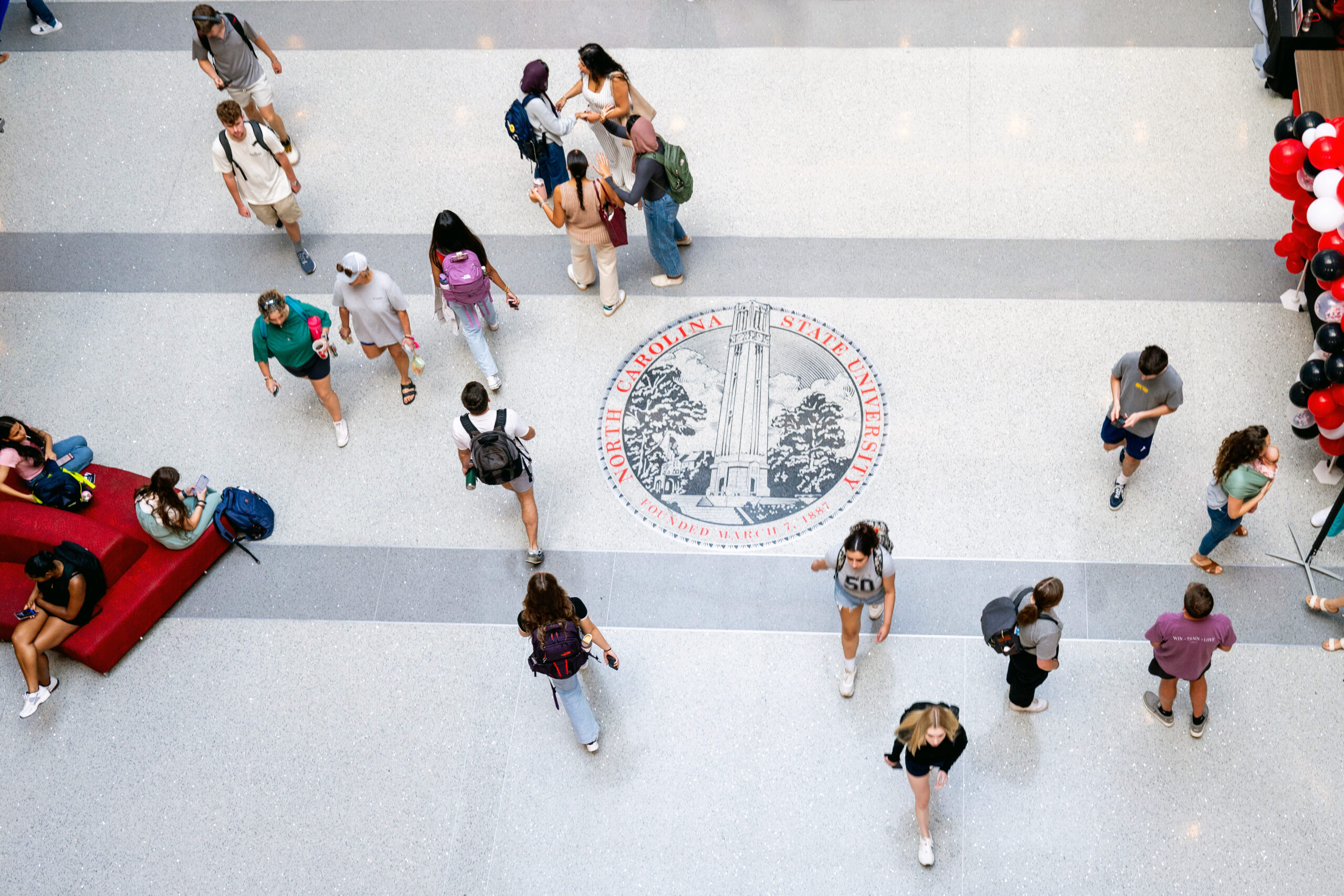 Photographed from above, students walk across the brightly lit floor of Talley Student Union, past the NC State crest displayed on the ground. 